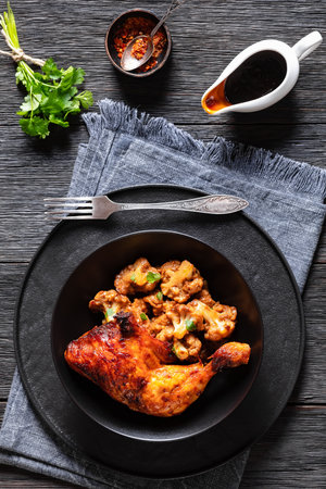 baked chicken leg with spicy roast cauliflower florets and fresh coriander on black plate on dark wooden table, vertical view from aboveの写真素材