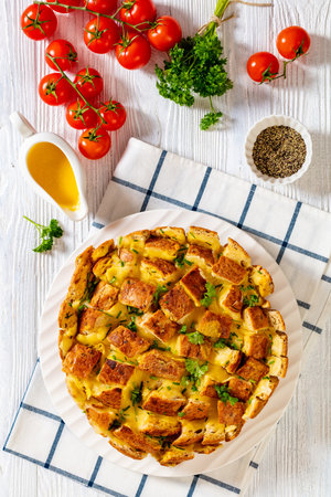 cheesy garlic bread, pull apart whole round bread on platter on white wood table with melted butter, tomatoes, and spices at background, vertical view from aboveの写真素材