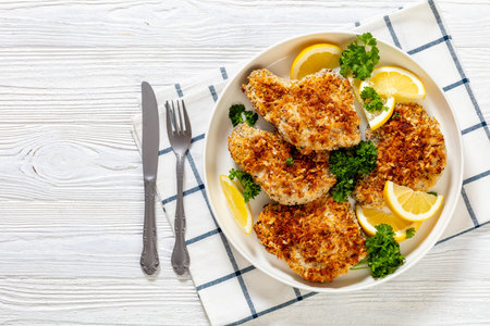 baked in oven panko breaded pork chops on bone with lemon slices and fresh parsley on plate on white textured wood table, horizontal view from above, flat lay, copy spaceの写真素材