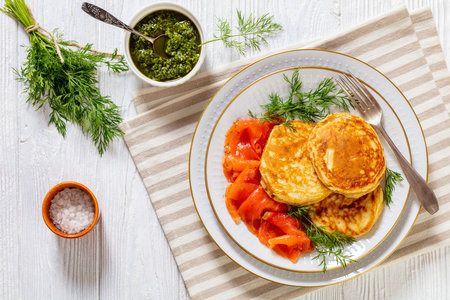 breakfast yeast pancakes with sliced cold smoked salmon and fresh dill on white plate on white wood table with sauce pesto on background, horizontal view from above, flat layの写真素材