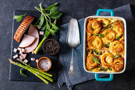 asparagus ham casserole topped with sponge cake circles in baking dish on concrete table with ingredients, horizontal view from above, flat layの写真素材
