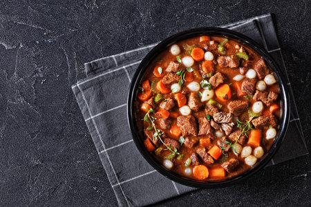 boneless beef short ribs stew with carrots and pearl onions in black bowl on concrete table with napkin, horizontal view from above, flat lay, free spaceの写真素材