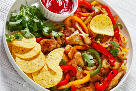 close-up of chicken fajitas of fried chicken breast strips, onion, green, yellow and red bell peppers on plate with corn tacos chipsの写真素材