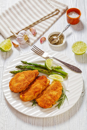 breaded chicken breasts with roast asparagus, rosemary and lime on white plate on white wooden table, vertical view from aboveの写真素材
