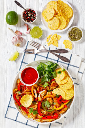 chicken fajitas of fried chicken breast strips, onion, julienned green, yellow and red bell peppers on plate with corn chips on white wood table with lime and seasoning, vertical viewの写真素材