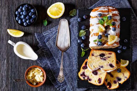 Lemon Blueberry Loaf with Lemon Glaze on slate board on dark wood table with fresh blueberries and cake shovel, horizontal view from above, flat layの写真素材