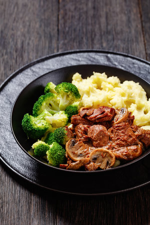 chopped beef steak mushrooms stew served with mashed potato and cooked broccoli florets in black bowl on dark wood table, vertical view, close-upの写真素材