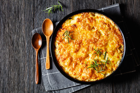 easy cheesy potato bake or potato casserole in black baking dish with, wooden spoons on dark wood table, horizontal view from above, flat lay, free spaceの写真素材
