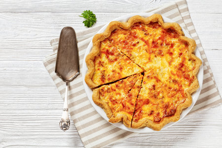 fluffy breakfast quiche with cheese and bacon filling on white plate on white wood table with cake shovel, horizontal view from above, flat lay, free spaceの写真素材