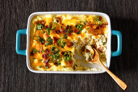 Loaded smashed potatoes with cheddar cheese, cream cheese, bacon, and cream in baking dish with wood spoon, flat lay, close-upの写真素材