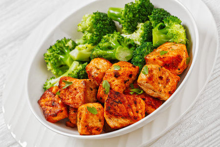 broiled salmon chunks with steamed broccoli florets in white bowl on white wood table, close-upの写真素材