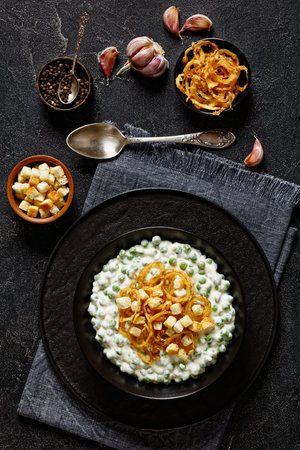 creamed peas topped with crunchy French fried onions and croutons in black bowl on concrete table with spoon and ingredients, vertical viewの写真素材