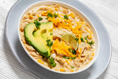 white chicken chili topped with shredded cheddar cheese, fresh slices of avocado and lime in white bowl on white wooden table, Dutch angle viewの写真素材