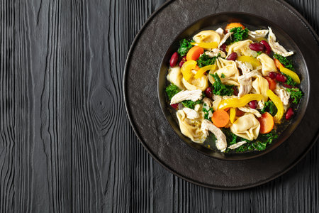 chicken minestrone soup with tortelloni and red kidney beans, kale and yellow bell pepper in black bowl on black wooden table, horizontal view from above, flat lay, free space, close-upの写真素材