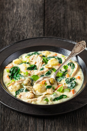 gnocchi zuppa toscana, Italian creamy chicken soup with gnocchi, spinach and vegetables in black bowl with spoon on dark wooden table, vertical view from above, close-upの写真素材