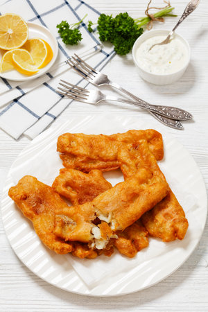 deep fried crispy beer batter cod fish fillet on white plate on white wooden table with forks and tartar sauce, vertical view from aboveの写真素材