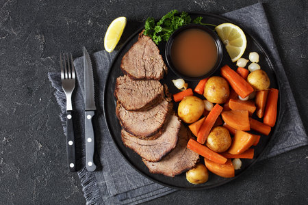 roast beef slices with dipping sauce and winter vegetables, potato, carrots, pearl onions, and rutabaga on black plate on concrete table with cutlery, horizontal view from above, free space, flat layの写真素材