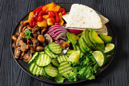 vegan fajitas, mushrooms, bell pepper, red onion, cucumber, avocado, cilantro, tortillas on black plate on black wooden table, landscape viewの写真素材