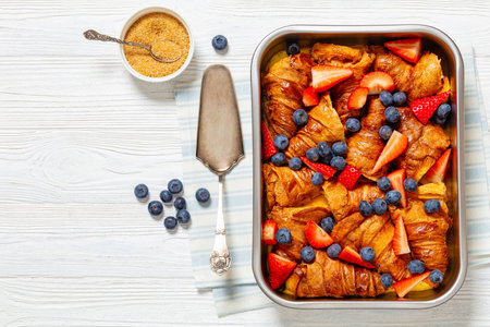 strawberry blueberry croissant casserole in baking dish on white wooden table with sugar and cake shovel, horizontal view from above, flat lay, free spaceの写真素材