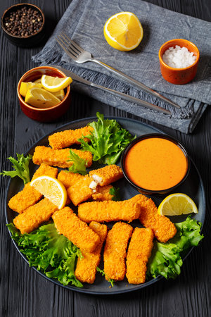crispy fried breaded fish sticks from white fish fillet served with fresh lettuce lemon wedges and hot sauce on platter on black wooden table, vertical viewの写真素材
