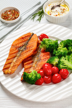 grilled salmon steaks with boiled broccoli and fresh cherry tomatoes on white plate on white wooden table with fork, seasoning, yogurt sauce based, vertical view, close-upの写真素材