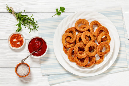 deep fried crispy onion rings on white plate on white wooden table with tomato sauce, seasoning and salt, horizontal view from above, flat layの写真素材