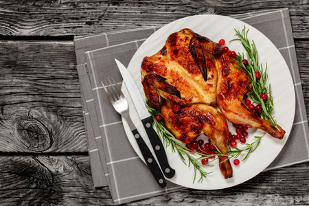 butterflied roast whole chicken on plate with rosemary and cranberry, on gray wooden rustic table with cutlery, horizontal view from above, flat lay, free spaceの写真素材