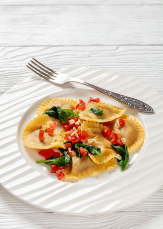 italian pasta mezzelune with tomatoes, garlic and basil on white plate with fork on white wooden table, vertical view from above, close-up, free spaceの写真素材