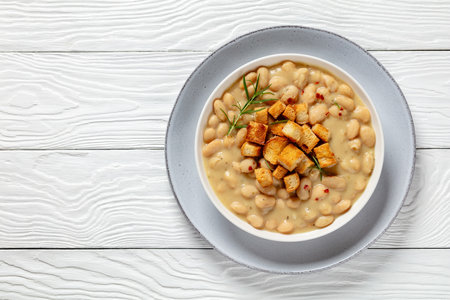 rosemary garlic creamy white bean soup topped with homemade croutons in white bowl on white wooden table, horizontal view from above, flat lay, free spaceの写真素材