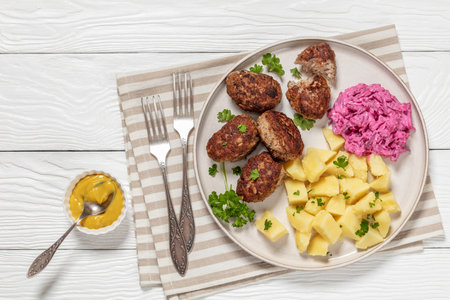 frikadeller, danish meatballs of minced pork with boiled potatoes and beetroot salad on plate on white wooden table with yellow mustard, horizontal view from above, flat lay, free spaceの写真素材