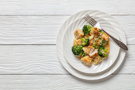 portion of Chicken Divan, succulent chicken bites in creamy sauce with broccoli topped with panko on white plate with fork on white wooden table, American cuisine, flat lay, free spaceの写真素材