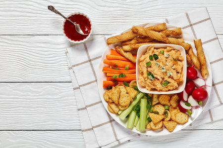 buffalo chicken dip with crackers, red radishes, carrot and celery sticks, grissini breadsticks on white platter on white wooden table, horizontal view from above, flat lay, free spaceの写真素材