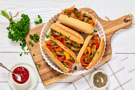 sausage, pepper, mushroom and onion hoagie sandwiches on wooden cutting board on white wooden table with tomato sauce and parsley, horizontal view from above, flat layの写真素材