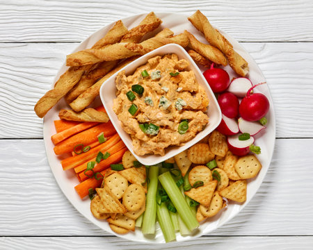 buffalo chicken dip with crackers, red radishes, carrot and celery sticks, grissini breadsticks on white platter on white wooden table, horizontal view from above, flat lay, close-upの写真素材