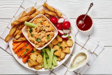 buffalo chicken dip with crackers, red radishes, carrot and celery sticks, grissini breadsticks on white plate on white wooden table, horizontal view from above, flat lay, close-upの写真素材