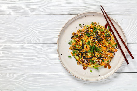 vegetarian okonomiyaki, Japanese savory pancake with cabbage, drizzle with sriracha mayo sprinkled with dried nori, sesame seeds, scallions on plate on white wooden table, flat lay, free spaceの写真素材