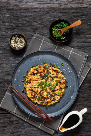 vegetarian okonomiyaki, Japanese savory cabbage hashbrown, drizzle with sriracha mayo sprinkled with nori, sesame seeds, scallions on plate on dark wooden table with ingredients, vertical viewの写真素材