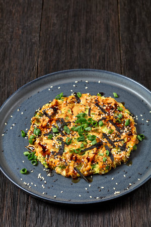 okonomiyaki, Japanese savory cabbage hashbrown, drizzle with sriracha mayo sprinkled with nori, sesame seeds, scallions on plate on dark wooden table, vertical view, close-up, free spaceの写真素材