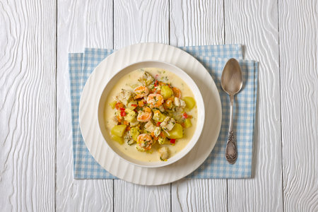 seafood chowder of shrimps, white fish, cream, broth and vegetables in white bowl on white wooden table with spoon, horizontal view from above, flat lay, free spaceの写真素材
