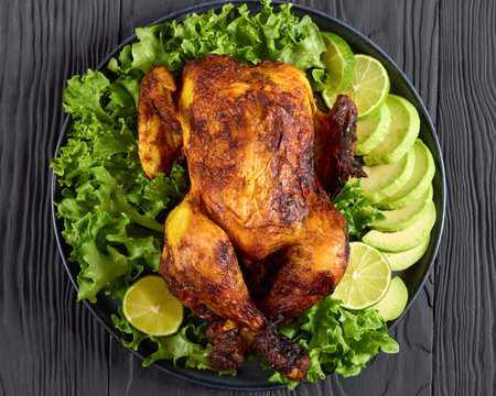 close-up of whole roasted chicken served with lettuce, avocado slices and lime on dark grey plate on black wooden table, horizontal view from above, flat layの写真素材
