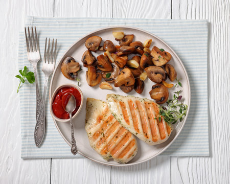 juicy grilled chicken breasts with sauteed garlic mushrooms on plate on white wooden table with forks, horizontal view from above, flat lay, close-upの写真素材