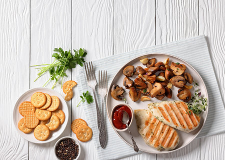 juicy grilled chicken breasts with sauteed garlic mushrooms on plate on white wooden table with crackers, tomato sauce and forks, horizontal view from above, flat lay, free spaceの写真素材