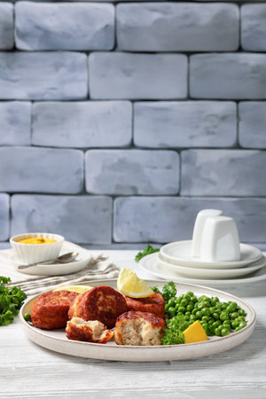 Danish atlantic cod fish cakes, fiskefrikadeller with green peas and lemon slices on a plate with cutlery on white wooden table with mustard and brick wall in background, vertical viewの写真素材