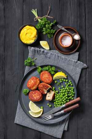 Danish atlantic cod fish cakes, fiskefrikadeller with green peas and lemon slices on a plate with cutlery on black wooden table with mustard, vertical view from aboveの写真素材