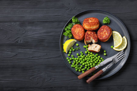 Atlantic cod fish cakes with green peas and lemon slices on a plate with cutlery on black wooden table, horizontal view from above, flat lay, free spaceの写真素材