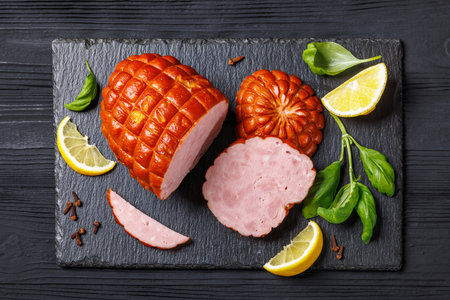 boneless smoked holiday ham on black stone board with pickles, cloves, basil and lemon on black wooden table, horizontal view from above, flat lay, close-upの写真素材