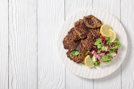 pork liver patties with fresh mixed salad and lemon slices on a white plate on white wooden table,...