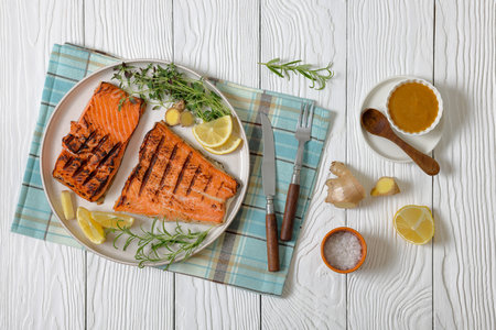 grilled salmon fish fillet on plate with ginger, rosemary, thyme, lemon on white wooden table with rustic cutlery and mustard, horizontal view from above, flat layの写真素材