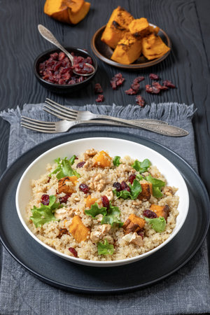 quinoa pumpkin salad with dried cranberries, walnuts  and fresh lettuce in white bowl on black wooden table with forks and ingredients, vertical view from aboveの写真素材