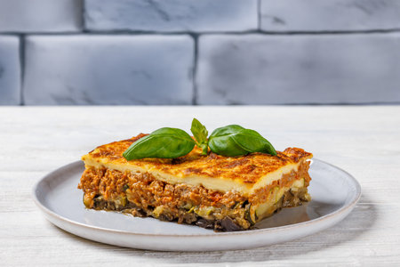 moussaka, layer of rich tomato beef sauce on fried eggplant topped with bechamel sauce on plate on white wooden table with brick wall in background, horizontal view from aboveの写真素材